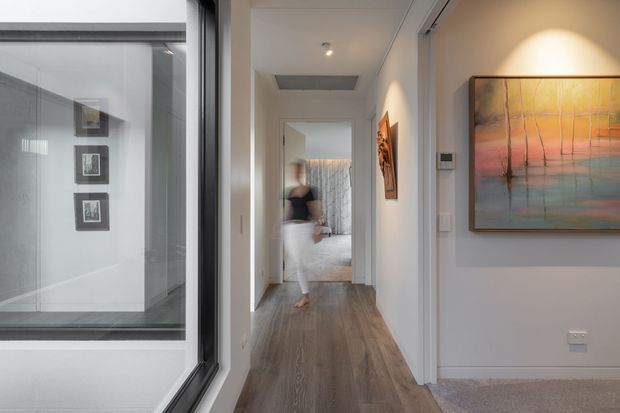 A bright contemporary residential hallway with grey-brown timber flooring, white walls, recessed ceiling lighting and a large colorful artwork. A glazed internal partition or window is visible on the left, while a bedroom or living space is visible at the end of the corridor.