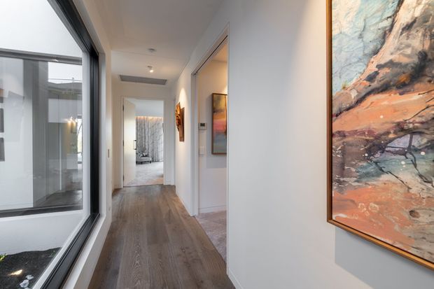 A bright contemporary residential hallway with warm timber flooring, white walls, recessed ceiling lights, and full-height glazing along one side overlooking an enclosed courtyard or lightwell. Framed abstract artwork adds colour to the corridor, with rooms opening off the hall and a softly furnished space visible at the far end.