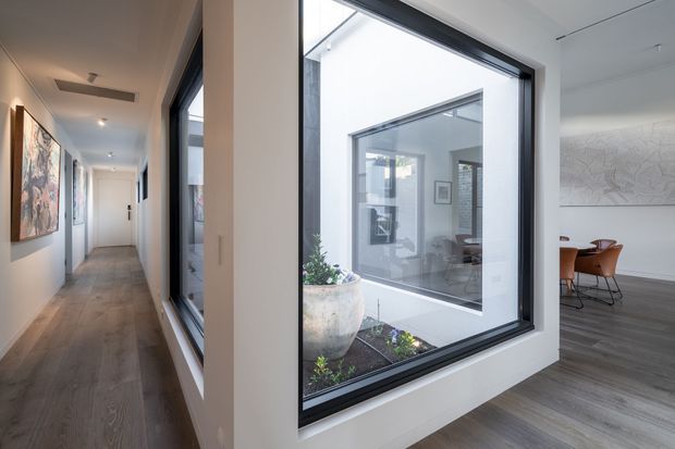 A bright contemporary interior showing a long hallway with white walls, large black-framed windows, and wide grey-toned timber flooring. The windows overlook an internal courtyard and provide views into an adjacent dining or living area furnished with a round table and brown leather chairs. Abstract artwork and a large wall artwork add visual interest, while the open, glazed arrangement creates a strong sense of connection between spaces.