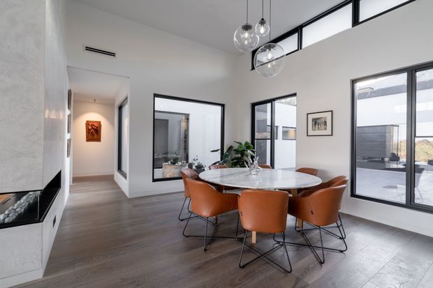 Bright contemporary dining area with a round marble-topped dining table and six rust-orange upholstered chairs, set against pale walls and dark timber-look flooring. Full-height glazing and high clerestory windows bring in abundant natural light, while a minimalist wall-mounted fireplace and glass pendant lights add refined architectural detail. An adjacent hallway and other interior spaces are visible through open doorways and glazing.