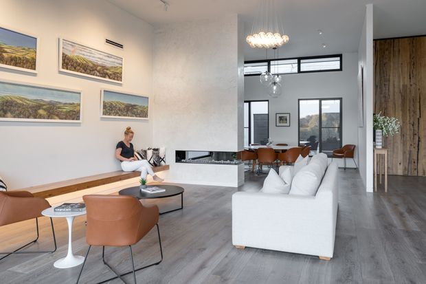 Bright open-plan contemporary living space with a white sectional sofa, brown leather lounge chairs, a low black coffee table, and a dining area beyond. A sculptural white fireplace divides the living and dining zones, while large dark-framed windows, polished concrete-style flooring, timber wall cladding, and globe pendant lights create a refined modern interior. Framed landscape artwork and built-in bench seating add warmth.