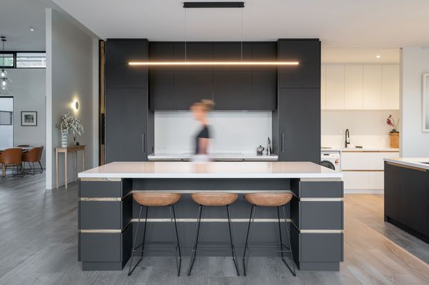 Contemporary open-plan kitchen centred on a large dark island with a pale benchtop and three timber bar stools. The kitchen features matte charcoal cabinetry, a white splashback, integrated appliances, and a linear pendant light. A lighter secondary kitchen area and dining space are visible on either side, with grey flooring throughout.