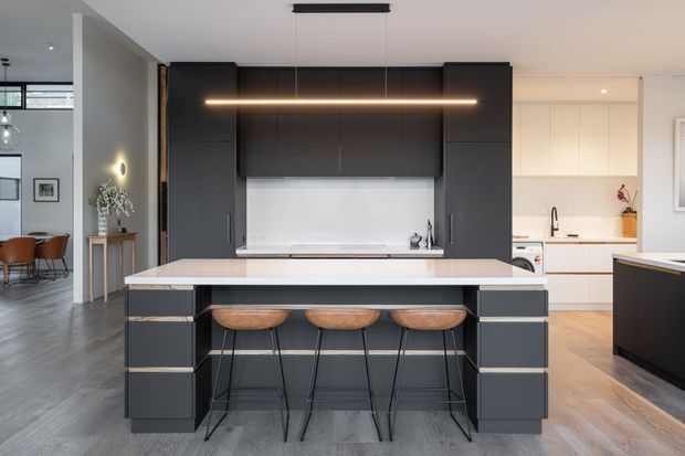Contemporary open-plan kitchen dominated by a large dark charcoal island with a pale stone benchtop and three timber bar stools. Full-height dark cabinetry frames a white splashback and concealed cooking area, while a lighter secondary kitchen zone is visible behind. Grey-toned flooring, minimalist linear pendant lighting, and clean integrated joinery create a refined, high-contrast interior.