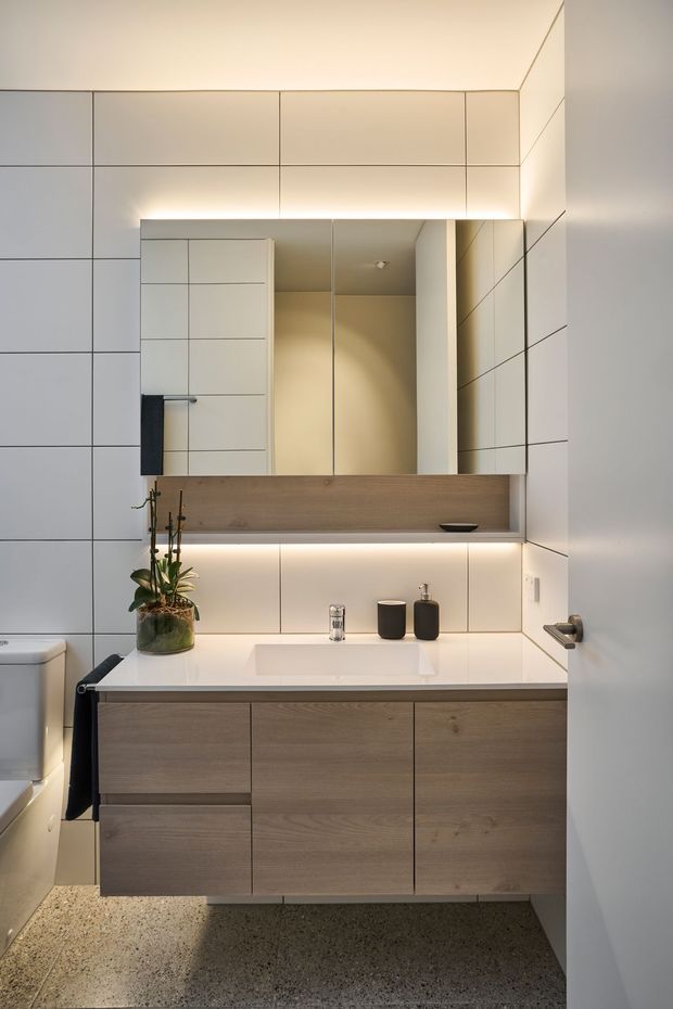 A compact contemporary bathroom featuring a floating timber-look vanity with a white integrated basin, large frameless mirror, and warm concealed LED lighting. The walls are clad in large light-grey rectangular tiles with crisp dark grout lines, complemented by a terrazzo-style floor. A toilet, black towel, small plant, and dark countertop accessories complete the restrained modern fit-out.