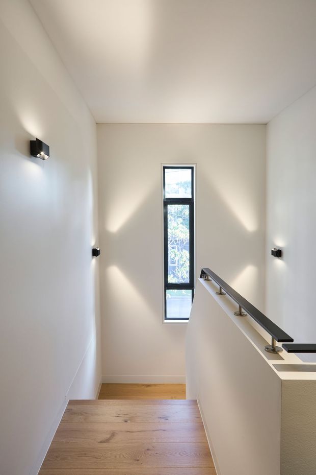 A minimalist stairway viewed from the upper landing, with broad light timber treads, smooth white walls, a slender black metal handrail, and a narrow vertical window bringing in natural light. Small black wall sconces create geometric pools of light along the stairwell.