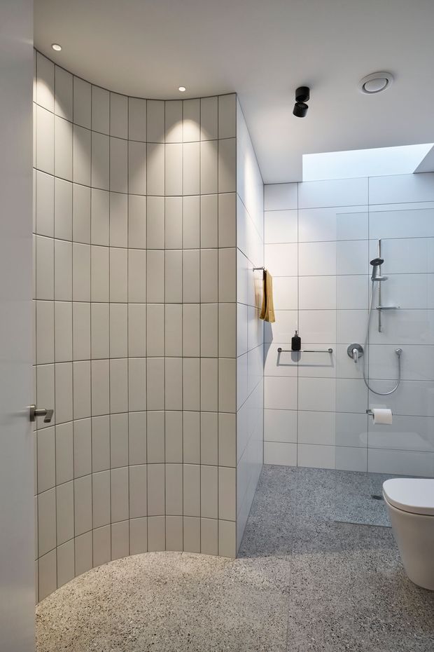 Contemporary bathroom with a curved tiled partition wall, large-format pale wall tiles, a walk-in shower, skylight, wall-mounted fixtures, toilet, and a speckled terrazzo-style floor. The space is bright, clean, and minimalist with subtle curved geometry.