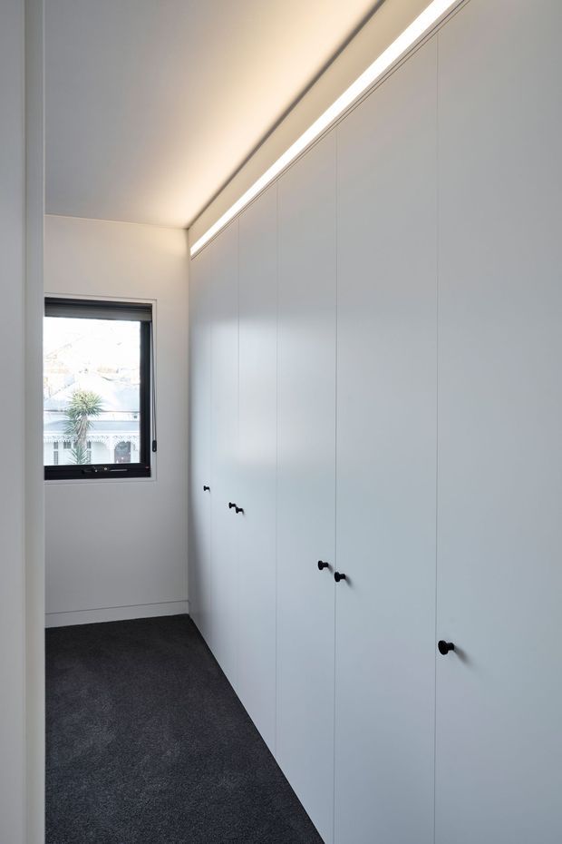 A narrow, minimalist storage corridor or walk-in wardrobe with full-height white built-in cabinetry along one wall, simple dark knobs, integrated linear ceiling lighting, white walls, and dark grey carpet. A small black-framed window provides daylight at the end of the space.