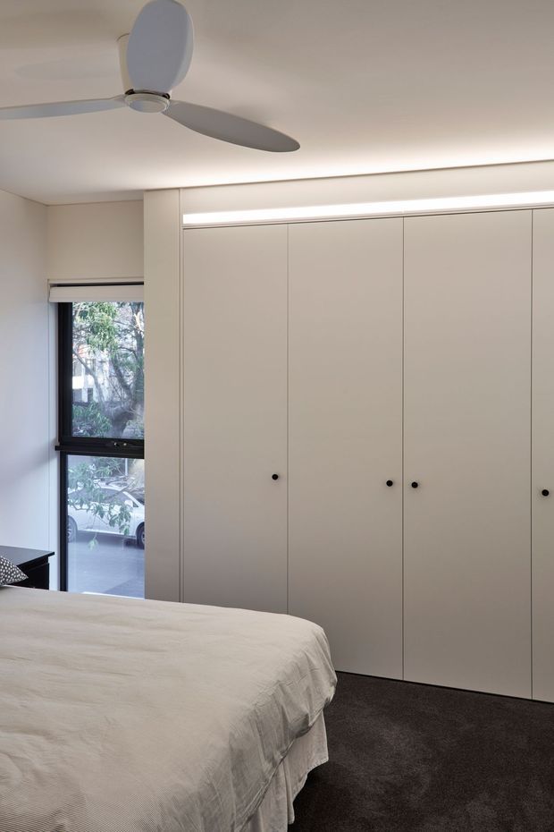 A minimalist bedroom with a large wall of full-height pale built-in wardrobes, small dark round pulls, dark carpet, and a low bed covered in light bedding. A black-framed window, recessed linear lighting, and a modern ceiling fan contribute to the clean, understated design.