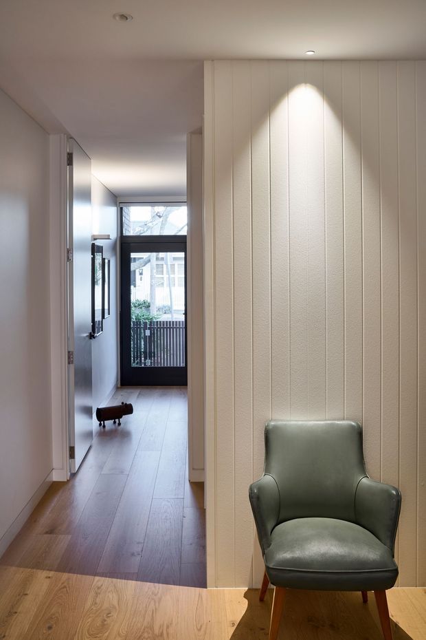 A warmly lit residential hallway or entry area with pale vertical wall panelling, a light timber floor, and a vintage green leather armchair in the foreground. The corridor leads toward a glazed exterior door with dark framing, while recessed ceiling lights and simple built-in cabinetry create a clean, refined interior.