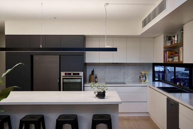 Contemporary kitchen with a large white island and three black stools, dark integrated cabinetry, stainless-steel appliances, pale flat-panel joinery, a grey stone-look splashback, timber flooring, and a timber open shelf displaying kitchen items. A window above the benchtop provides an urban outlook.