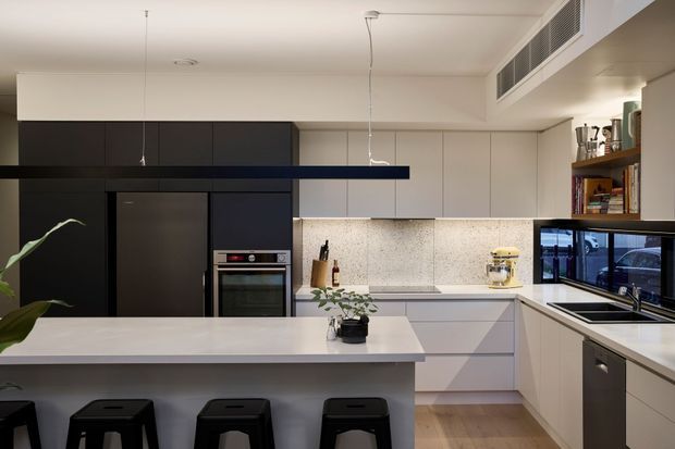 Contemporary open-plan kitchen with a large pale island and three black stools, matte dark cabinetry around the refrigerator and oven, light upper and lower cabinetry, a speckled stone backsplash and benchtops, integrated appliances, timber flooring, and open shelving. A window above the sink provides an exterior view, while a concealed linear air-conditioning grille runs along the ceiling edge.