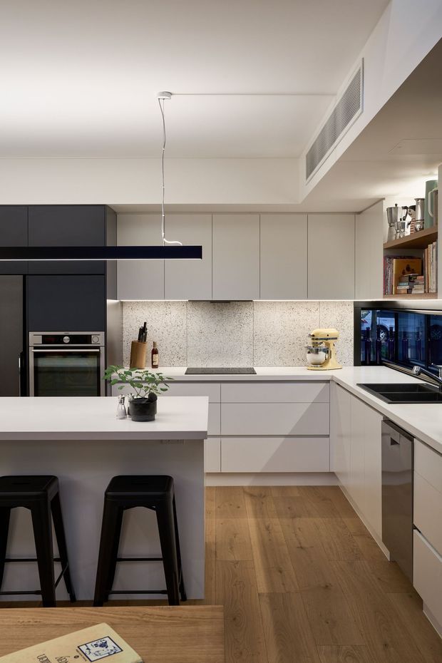 A contemporary open-plan kitchen with a matte white and dark charcoal cabinetry scheme. The space features an L-shaped kitchen with integrated appliances, pale stone-look splashback, white benchtops, a timber island with black bar stools, open shelving, and warm timber flooring. A slim black linear pendant light and under-cabinet lighting provide contrast.