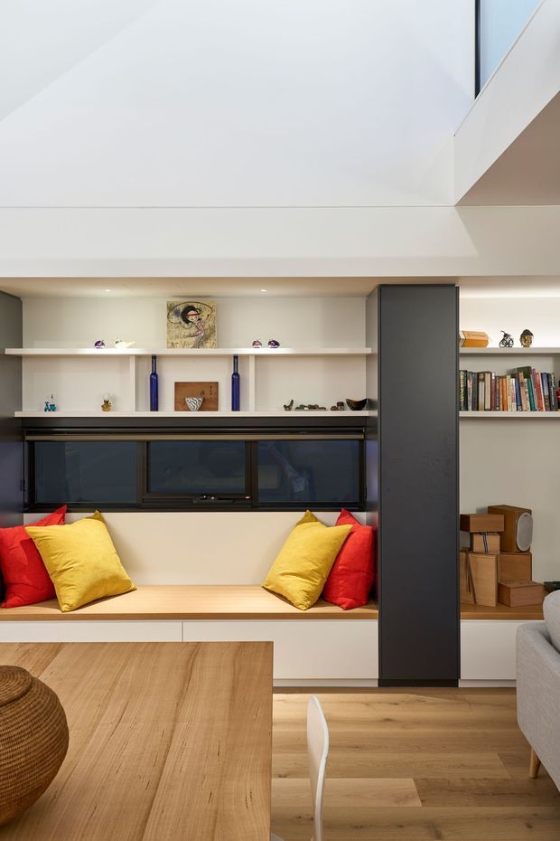 Bright contemporary living room with a built-in window seat, vivid red and yellow cushions, extensive open shelving, dark integrated cabinetry, timber flooring, and a large timber dining table. A dramatic white geometric ceiling and clerestory-like opening contribute to the architectural character.