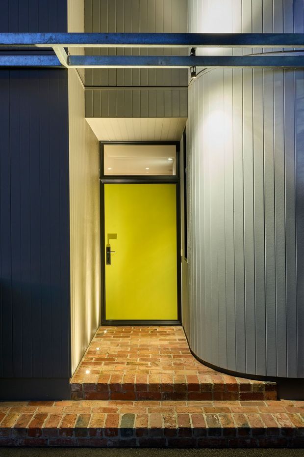 A narrow covered entrance alcove with a vivid yellow entry door, black framing, dark and grey vertical-clad walls, exposed blue-grey steel beams overhead, and a red-brown brick-paved threshold with steps. The image emphasises the strong colour contrast and layered architectural entry details.