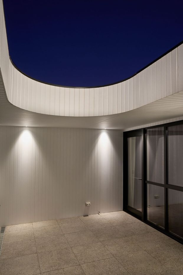 A sheltered contemporary courtyard or covered outdoor terrace with a dramatic curved white vertical-panel canopy and matching wall cladding. Large dark-framed glazed doors line one side, while recessed wall lighting illuminates a pale tiled floor and the curved ceiling form.