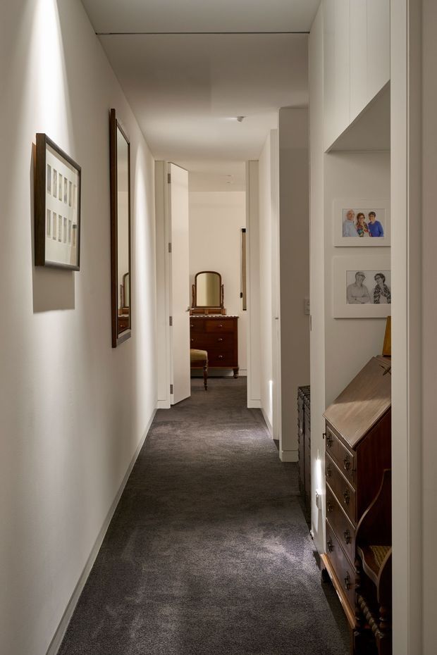 A long, narrow residential hallway with white painted walls, recessed wall niches displaying framed artwork and photographs, dark grey carpet, and white doors. A traditional dark timber dresser is visible in the foreground and another dresser with a mirror is visible at the end of the corridor. The space is softly lit and uncluttered, with a calm, neutral palette.
