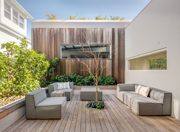 A bright contemporary courtyard terrace enclosed by residential buildings. The main feature is a compact timber-clad pavilion with a flat roof and horizontal window, opening onto a spacious timber deck furnished with grey outdoor lounge seating. Planted raised beds and a small tree soften the courtyard, while white rendered walls and large openings create a clean, private outdoor living space.