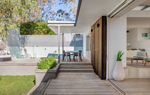 Bright contemporary courtyard and rear exterior of a modern home, viewed from a timber deck toward an outdoor dining area and pool. The open sliding glass doors reveal a refined living room with light neutral finishes and mid-century-influenced furniture. The architecture features clean horizontal lines, a flat roof, extensive glazing, timber cladding, concrete steps, and landscaped raised planters.