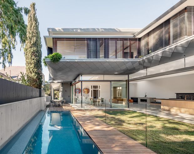 Contemporary multi-level residence viewed from a landscaped courtyard, featuring a narrow swimming pool, timber deck, extensive glazing, a covered outdoor living area with built-in cabinetry, and an upper-level cantilevered form clad in faceted grey panels. Horizontal timber or metal sun-shading screens span the upper windows, with clean flat rooflines and integrated planting.
