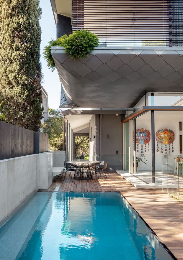 A contemporary multi-level residential exterior viewed along a narrow backyard pool. The scene features a cantilevered upper floor with a distinctive diamond-patterned grey underside and fascia, horizontal timber sun-louvres, glass balustrades, and a glazed ground-floor living area. A timber pool deck, outdoor dining setting, boundary wall, mature vegetation, and vivid blue swimming pool create a refined urban courtyard setting.