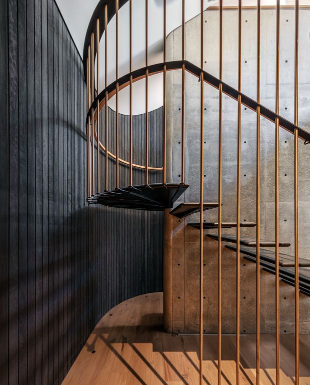 A sculptural curved staircase viewed from within a tall stairwell. The stair has dark timber or metal treads, slender warm copper-toned vertical balusters, and curved dark handrails. The surrounding walls combine vertically ribbed dark cladding with exposed concrete panels, while warm timber flooring and stair elements add contrast. Strong natural light creates linear shadows across the floor.