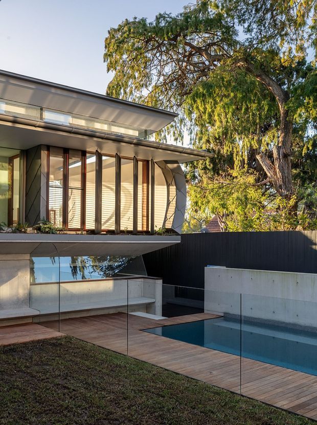 Exterior view of a contemporary multi-level home with broad roof overhangs, extensive glazing, timber-framed vertical windows, and faceted structural detailing. A timber deck surrounds a compact swimming pool, enclosed by frameless glass balustrades and dark privacy fencing. Mature trees frame the property.