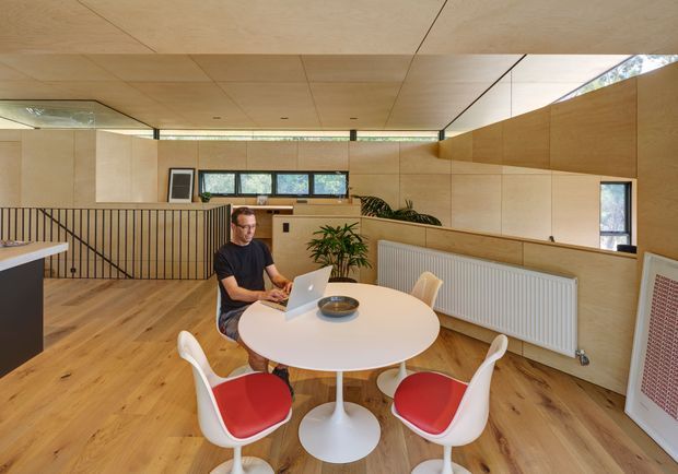 A warm, compact open-plan interior with a round white dining table and contemporary white chairs with red seat cushions in the foreground. A person is seated at the table using a laptop. The space includes light timber flooring, extensive pale timber wall and ceiling panelling, built-in timber cabinetry, a linear radiator, a compact kitchen or work area beyond a black vertical-bar balustrade, and generous horizontal windows. A large mirror reflects the room, while plants and artwork add small decorative touches.