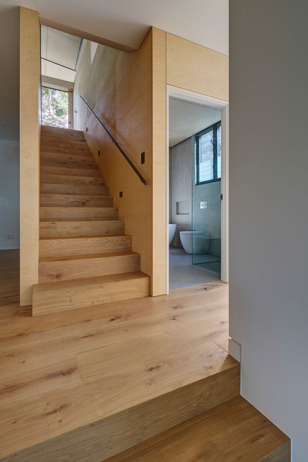 Interior view of a compact, warm-toned stairway with broad solid timber treads and matching timber flooring. Pale plywood or timber-lined walls frame the staircase, with a slim dark metal handrail. A bathroom with a toilet, basin and shower area is visible through an open doorway beside the stairs. Natural light enters through upper glazing and a small window, creating a calm, contemporary architectural interior.