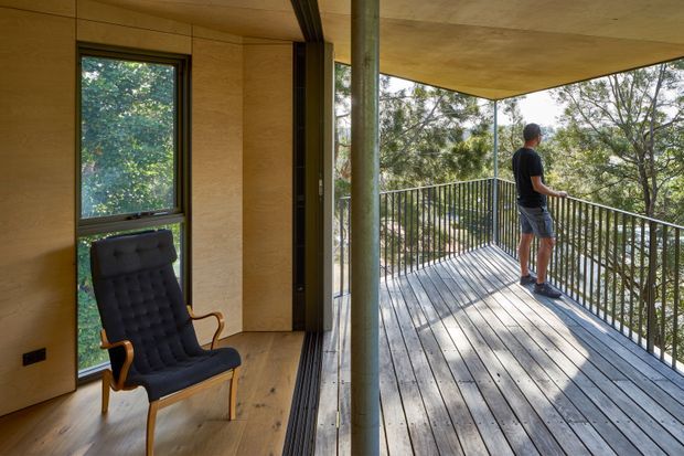 A warm, timber-lined interior opens directly onto a spacious elevated deck through fully retractable glazing. The room contains a dark upholstered lounge chair, while the deck has weathered timber boards, slender metal balustrades, and a central structural post. Dense trees surround the outdoor platform, creating a secluded connection to nature.