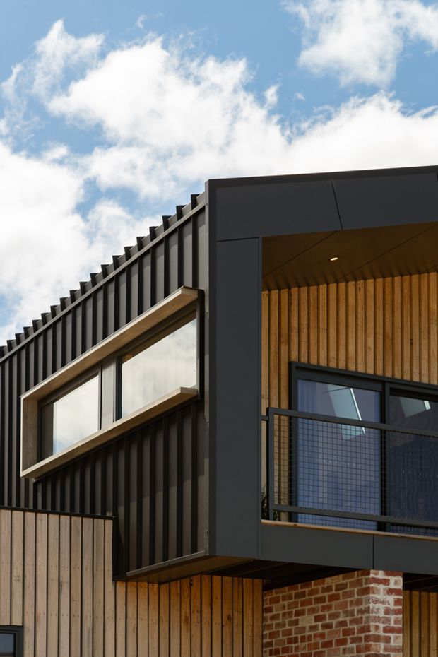 Close-up exterior architectural view of a contemporary multi-level building. The facade combines dark vertical metal cladding, warm vertical timber lining and fencing, large dark-framed windows, a projecting balcony with black mesh balustrade, and a brick base. The image emphasizes strong rectilinear forms and a partially recessed, timber-lined upper-level opening beneath a flat roofline.