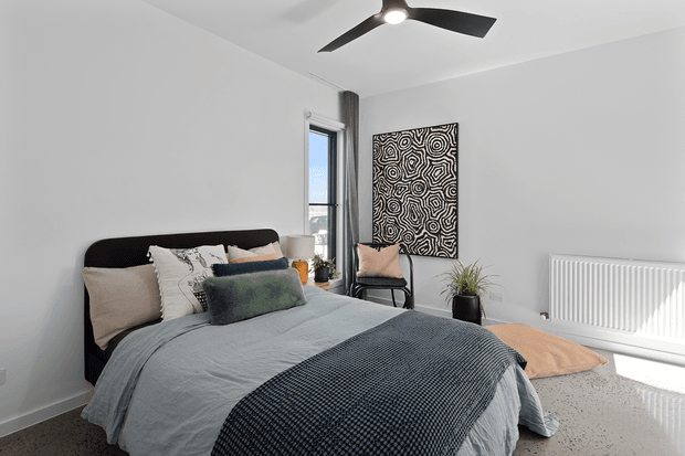 Bright contemporary bedroom with a low upholstered dark bed, layered neutral and muted green bedding, a textured dark throw, a black ceiling fan, white walls, a tall window, patterned wall art, a potted plant, and a white panel radiator. The room has a calm, restrained palette and polished concrete-style flooring.