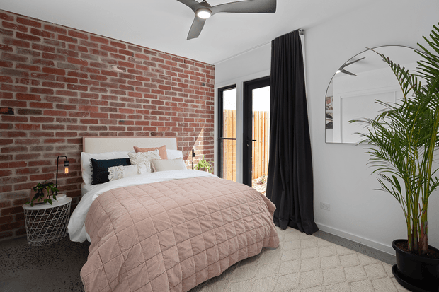 A bright, contemporary bedroom with a low-profile upholstered bed dressed in layered white and blush bedding. The room features an exposed red brick feature wall, black-framed glazed doors with full-height dark curtains, a pale grey painted wall, a large arched mirror, a ceiling fan, a textured light rug, and a tall potted palm.