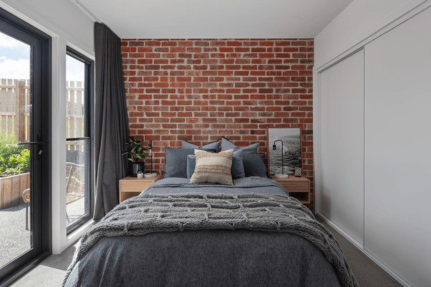 A compact, contemporary bedroom with a bed positioned against an exposed red brick feature wall. The room includes charcoal and grey bedding, layered cushions, a chunky knitted throw, dark curtains, timber bedside tables, potted plants, and a large white built-in wardrobe. Full-height glazed doors on the left open to a small outdoor area, bringing in natural light.