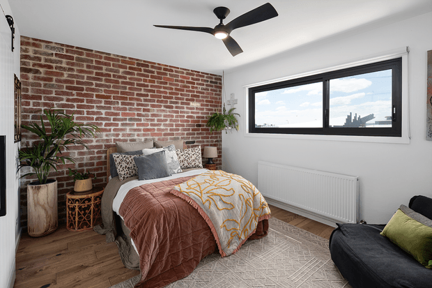A bright contemporary bedroom with a bed positioned against an exposed red brick feature wall. The room includes white walls, timber flooring, a large black-framed horizontal window, a ceiling fan, bedside tables, indoor plants, patterned bedding, and a dark sofa or lounge chair in the foreground. The styling combines industrial architectural detailing with warm, contemporary furnishings.