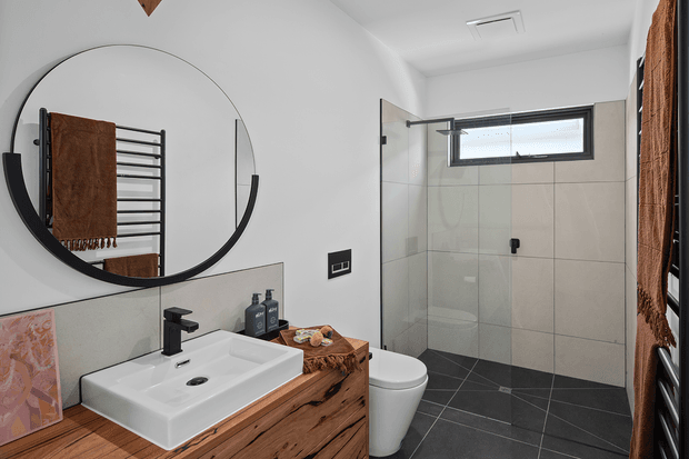 A contemporary bathroom featuring a wall-mounted timber vanity with a white rectangular basin, matte black tapware, a large round black-framed mirror, a wall-hung towel warmer, and a frameless glass shower enclosure. The shower has large light-grey wall tiles, dark charcoal floor tiles, and a high horizontal window. Earthy timber and brown textiles add warmth to the monochrome palette.