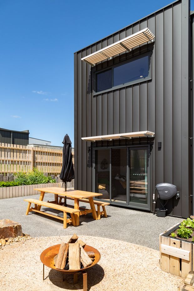 A sunny outdoor courtyard beside a modern two-storey building clad in dark vertical metal panels. The space includes a timber picnic table with benches, a covered barbecue, raised timber garden planters, a gravel and concrete-paved surface, and a circular outdoor fire pit with stacked firewood. Large glazed doors and windows connect the courtyard to the building interior, with timber sunshades above the openings.
