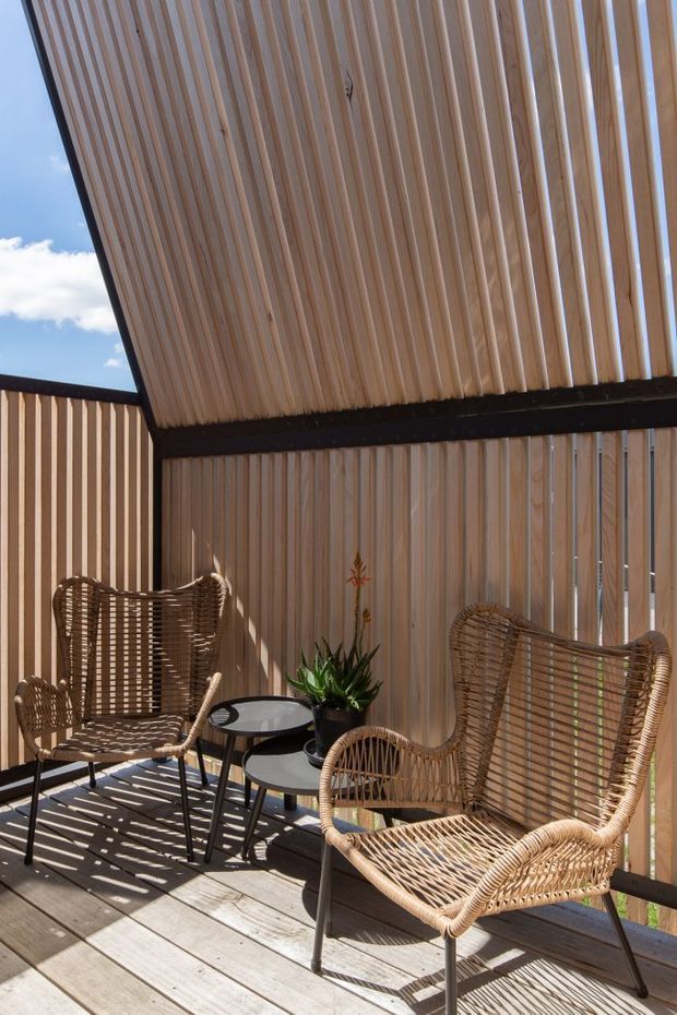 A sunlit outdoor terrace or balcony with timber decking, vertical timber screening, and a slatted timber canopy. Two woven rattan lounge chairs with black metal legs surround small round tables and a potted plant. Strong linear shadows create a warm, relaxed atmosphere.
