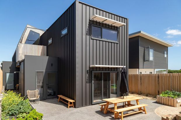 A sunny exterior view of a contemporary multi-storey residential building with a tall dark vertical-panel-clad central facade, large windows with external sunshades, balconies, and a ground-level glazed door. A timber-clad neighbouring dwelling, timber boundary fencing, paved courtyard, raised planter boxes, and picnic-style outdoor seating are visible.