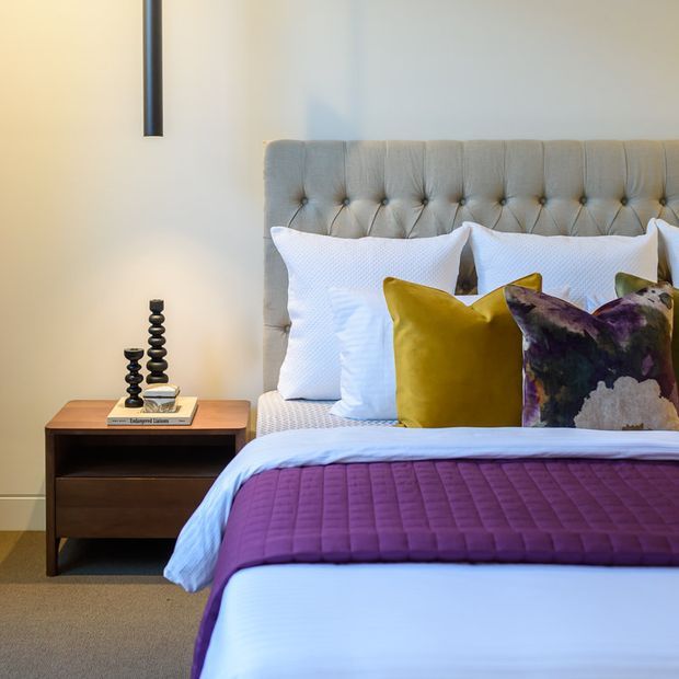 A stylish bedroom featuring a tall tufted upholstered headboard, white bedding, mustard and abstract-patterned accent cushions, and a deep purple quilt. A timber bedside table with decorative objects sits beside the bed, while a slim black wall light and neutral carpet complete the warm, contemporary setting.