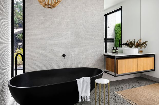 A refined contemporary bathroom featuring a freestanding matte black bathtub, textured light wall tiles, black-framed floor-to-ceiling glazing, and a floating timber vanity with a white countertop and integrated basin. Terrazzo-style flooring, a woven pendant light, and a neutral rug complete the warm monochromatic scheme. Garden planting is visible through the glazing.