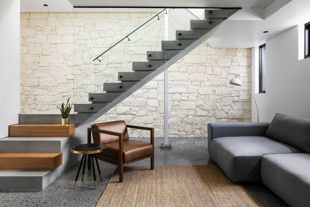 Contemporary living area beneath a minimalist floating staircase. A pale exposed-stone feature wall forms the backdrop, with grey concrete stair treads and flooring, clear glass balustrading, a dark grey sectional sofa, a leather lounge chair, timber stair details, and a natural woven rug. The space is bright and refined with strong textural contrast.
