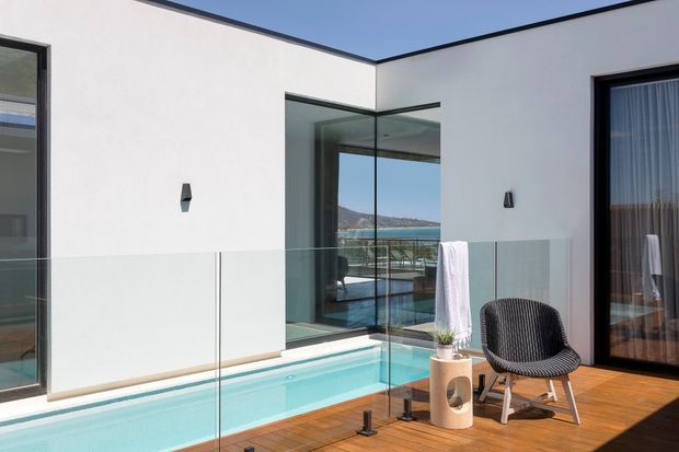 Modern white rendered courtyard terrace with a narrow turquoise swimming pool, frameless glass balustrades, timber decking, floor-to-ceiling glazing, and a black-framed glass corner opening. Outdoor seating and a small side table create a compact lounge area, with a coastal water view visible through the glazing.
