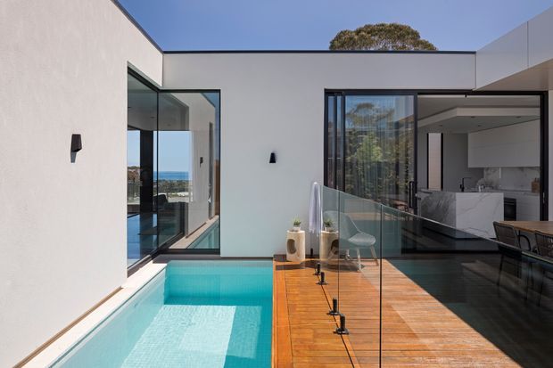 Modern courtyard-style residential exterior featuring a turquoise swimming pool alongside a warm timber deck, frameless glass balustrade, and crisp white rendered walls. Large black-framed glazed openings connect the courtyard to a contemporary kitchen and living area, with a glimpse of the coastal landscape beyond.
