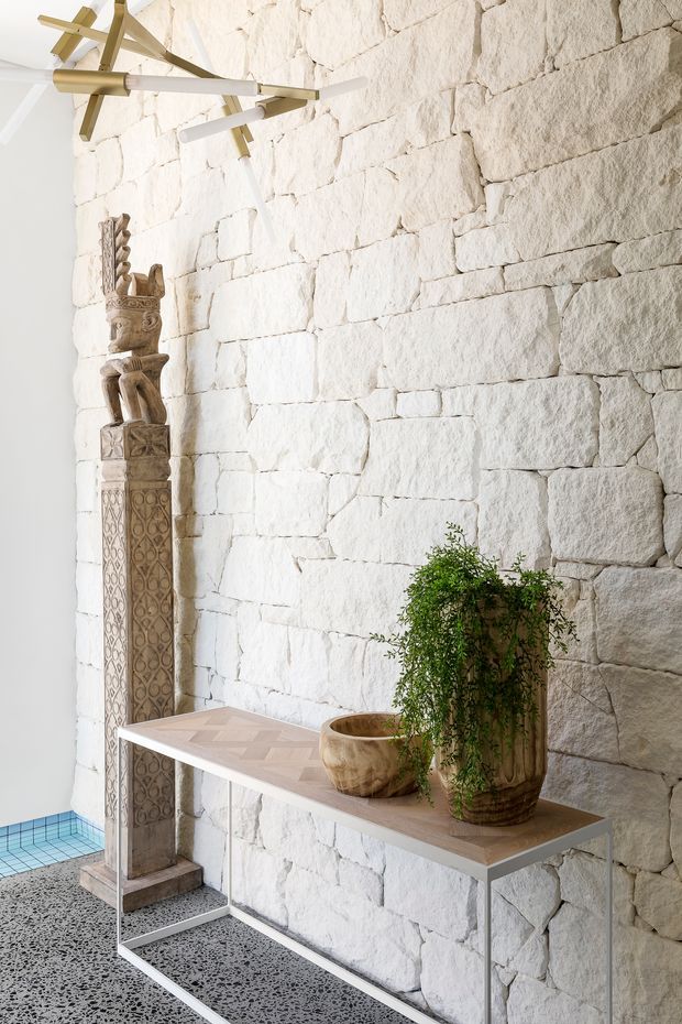 Bright, minimalist interior vignette featuring a white rough-cut stone masonry wall, a slim pale timber console table with slender metal legs, potted greenery, an ornate carved wooden sculpture, and a sculptural brass and white linear pendant light. A small glimpse of blue pool tiling appears at the lower left.
