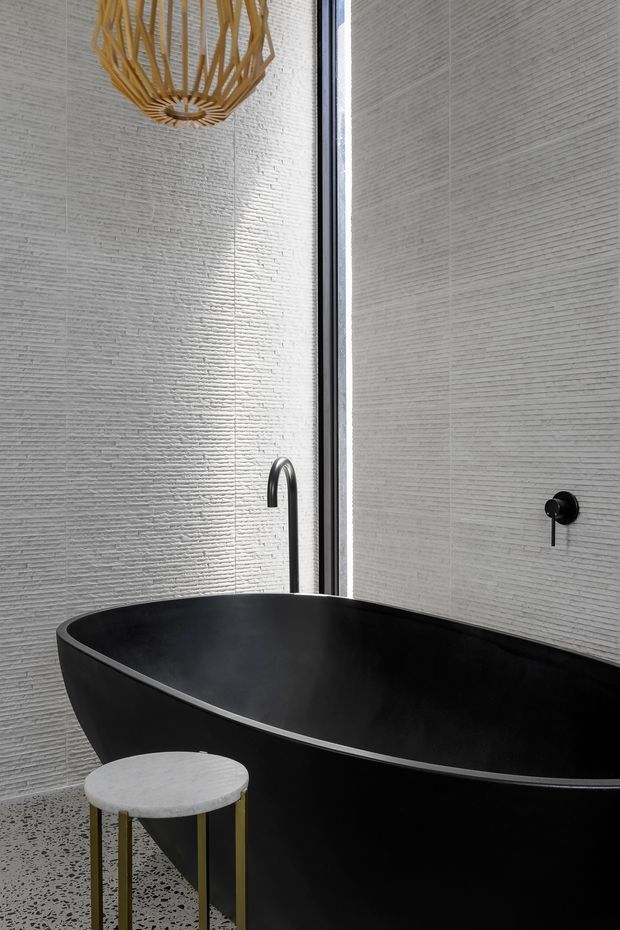 A refined modern bathroom featuring a large matte-black freestanding bathtub, a black wall-mounted tap, and a tall narrow mirror or glazed opening. The walls are clad in finely textured light-grey horizontal tile, while the floor appears to be terrazzo with dark aggregate. A small round light stone side table with slender brass-toned legs sits beside the tub, and a sculptural woven pendant adds warmth overhead.