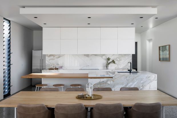 Contemporary open-plan kitchen and dining area with a large marble-look island, white handleless overhead cabinetry, integrated appliances, timber breakfast bar, and a long timber dining table with upholstered chairs. A layered ceiling with recessed downlights and a linear feature bulkhead adds definition, while full-height glazing and a framed artwork brighten the neutral interior.