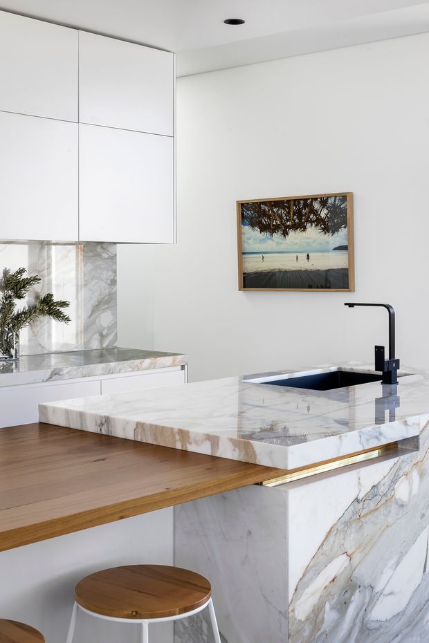 Bright contemporary kitchen featuring a large veined marble waterfall island and matching marble benchtops, crisp white handleless cabinetry, timber breakfast-bar extension, black undermount sink and matte black tapware. Warm timber flooring and stools contrast with the predominantly white scheme, while a framed beach photograph adds a small decorative accent.