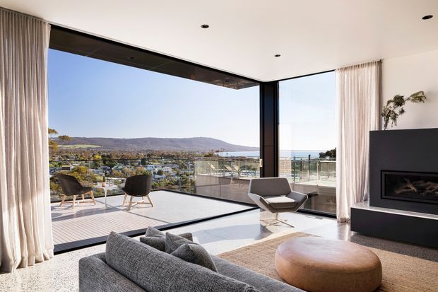 A refined contemporary living room with floor-to-ceiling corner glazing overlooking a broad landscape and water view. The room features a large grey sectional sofa, sculptural lounge chair, rounded timber coffee table, neutral textured rug, full-height curtains, recessed ceiling lights, and a striking dark built-in fireplace wall. An outdoor terrace with seating is visible beyond the glazing.