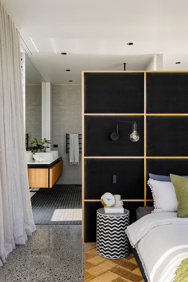 A contemporary open-plan bedroom and bathroom suite. The foreground features a bed with white bedding, green accent cushions, a patterned black-and-white bedside stool, and a black room divider with warm timber edging and integrated lighting. Behind it is a compact bathroom with a timber vanity, white basin, black benchtop, wall-mounted fixtures, a freestanding white bath, tiled walls and dark grid-pattern floor tiles. The space has polished concrete flooring, sheer curtains and recessed ceiling lights.