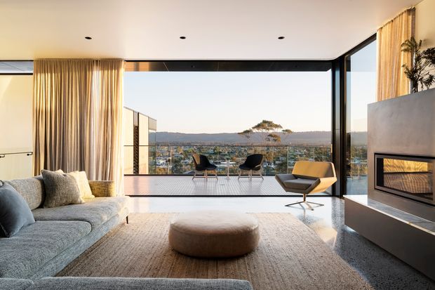 A refined contemporary living room with expansive floor-to-ceiling glazing opening onto a balcony and distant hillside view. The room features a large textured sectional sofa, neutral area rug and ottoman, a sculptural lounge chair, full-height curtains, polished reflective flooring, recessed ceiling lights, and a substantial built-in fireplace surround.